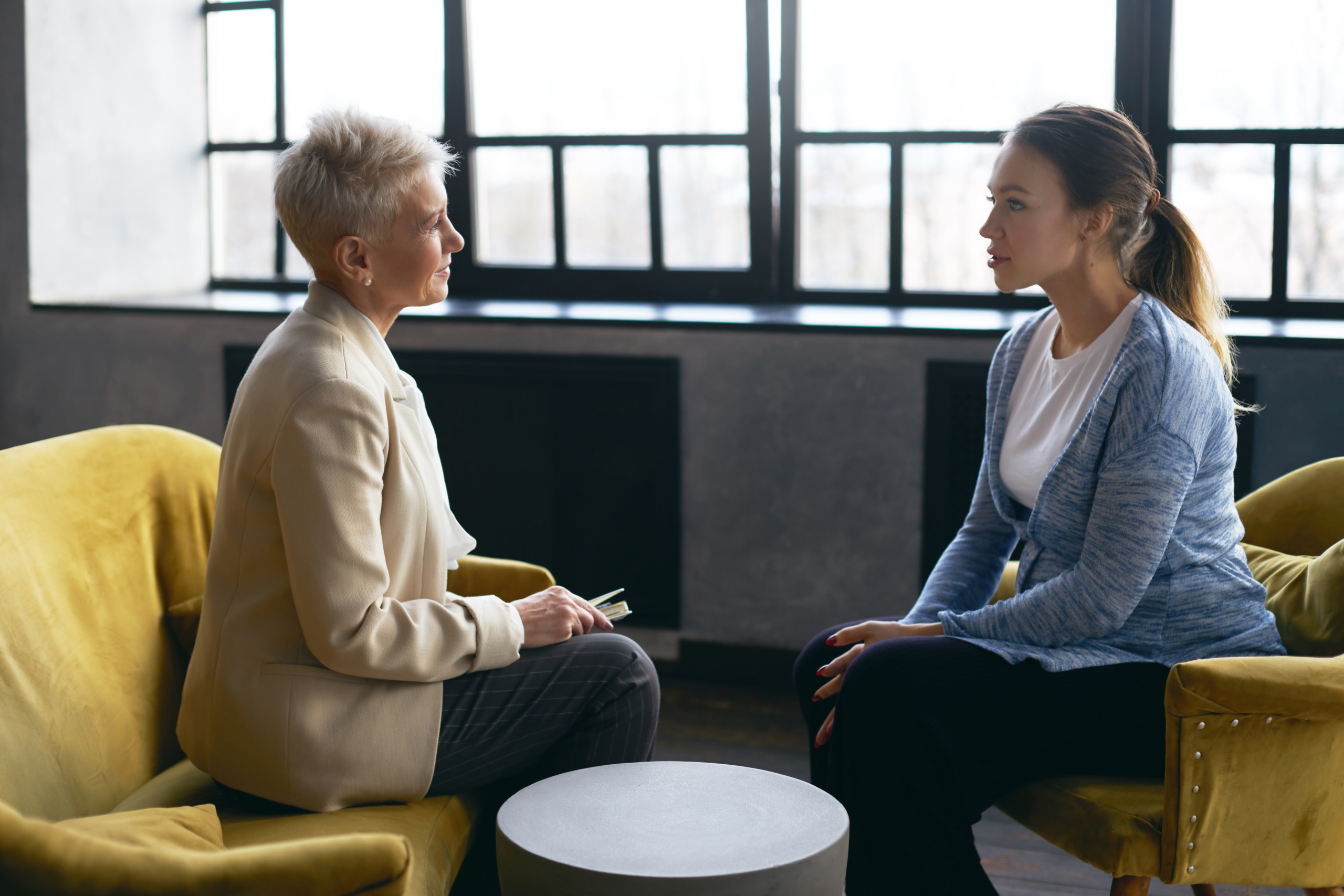 Side view image of middle aged female psychologist discusses the problems with young pregnant woman, sitting in armchairs, facing each other. Prenatal psychological therapy